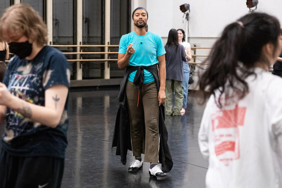 Person in blue shirt and dark pants walks in a rehearsal room full of dancers