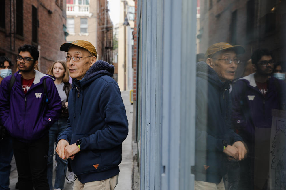 A man is reflected in a window in an alleyway, young people are seen to the side listening to him talk