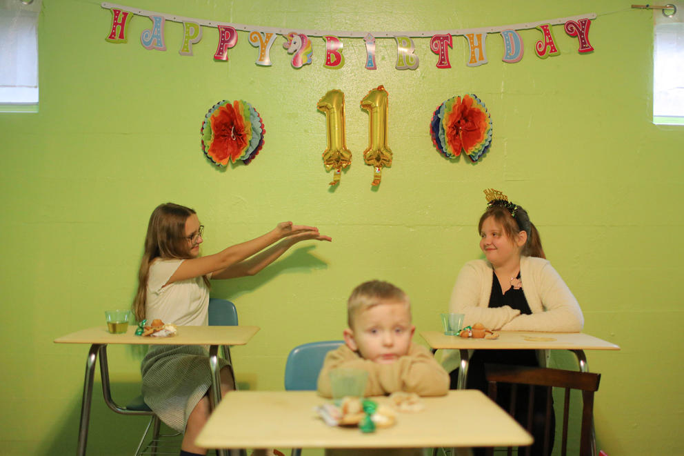 A hanging sign reads Happy Birthday 11 over two girls interacting. A young boy is in the foreground with his head on a desk.