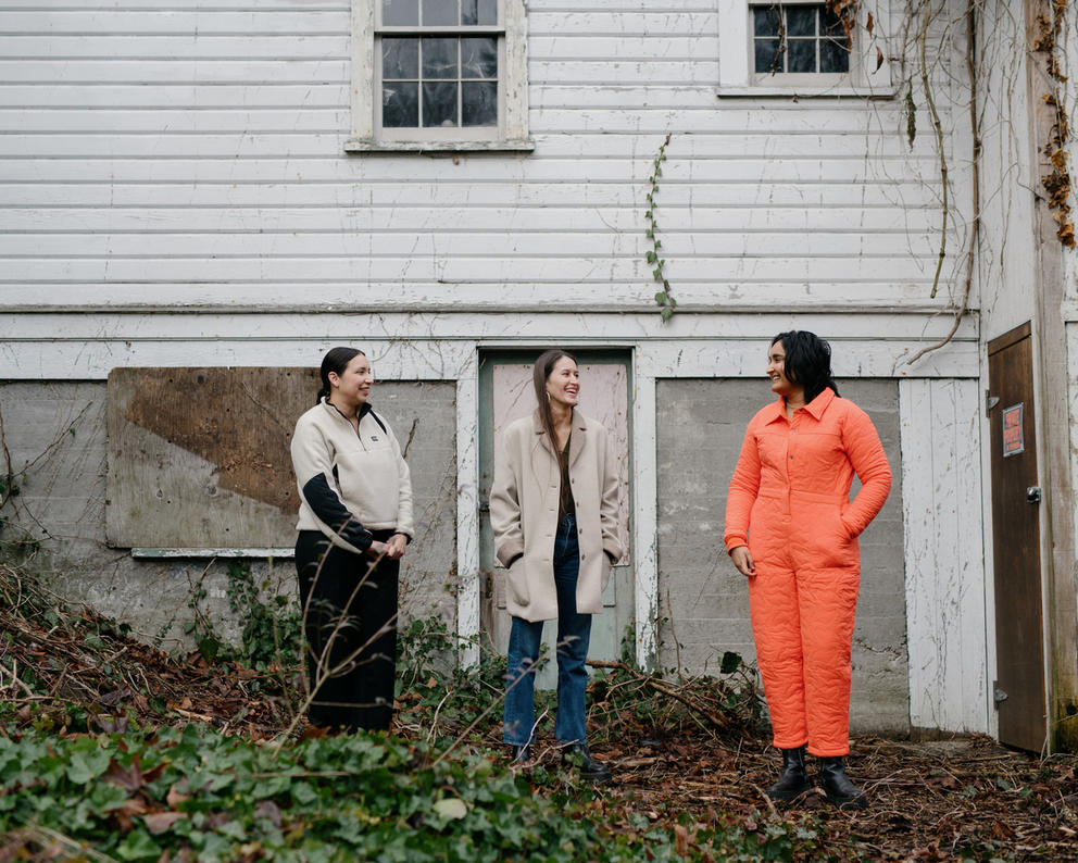Three people stand in front of a shed, one person wears a beige fleece, the other a beige vest and the third person an orange jumpsuit
