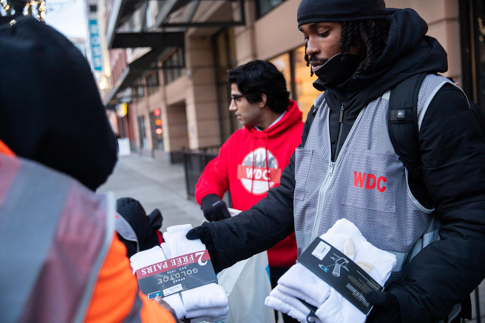two men hand out socks to people on the street in seattle. one is wearing a gray vest. the other is wearing a red sweatshirt.