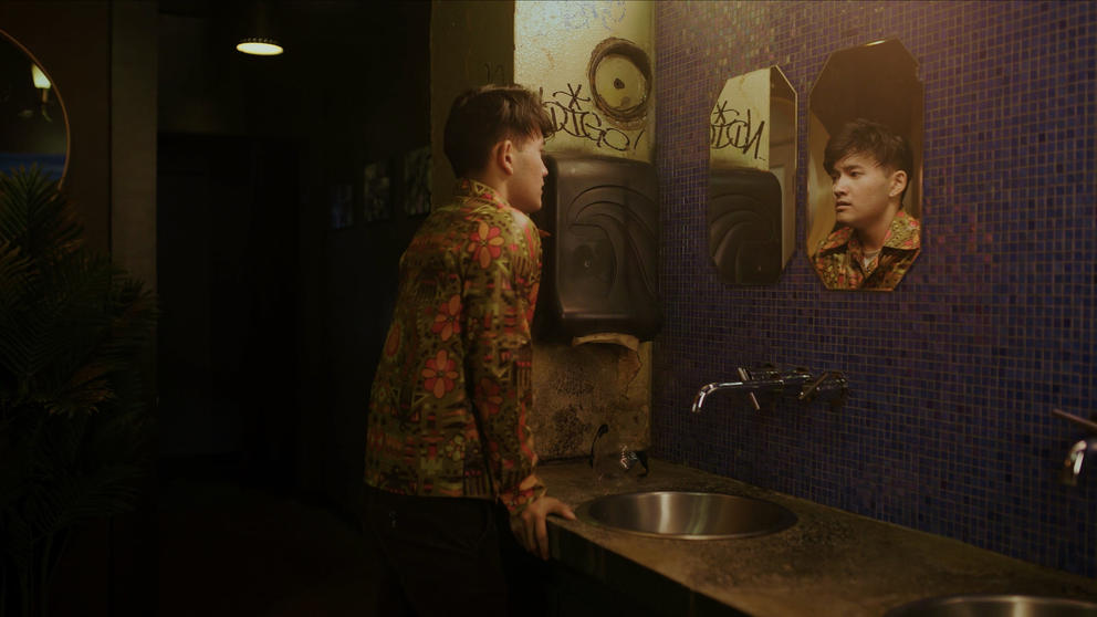 A wide shot of an Asian man staring at his reflection in the mirror pensively
