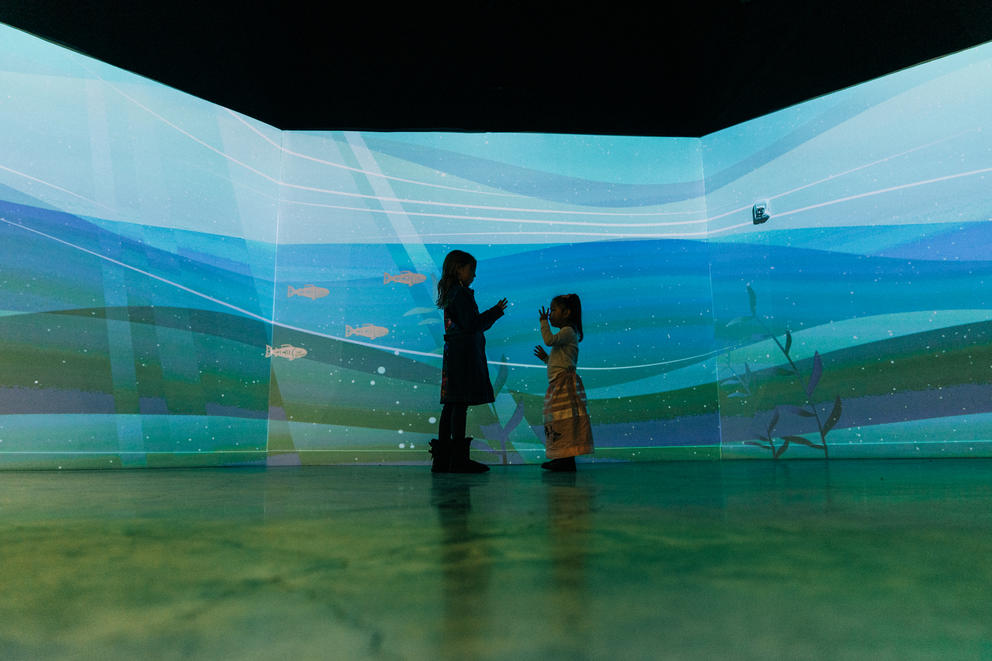 Two children stand in front of a projection of water with fish swimming