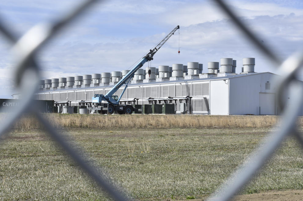 A crane is seen through a wire fence at a building holding computer equipment used in cryptocurrency "mining" that relies on electricity in Hardin, Montana.