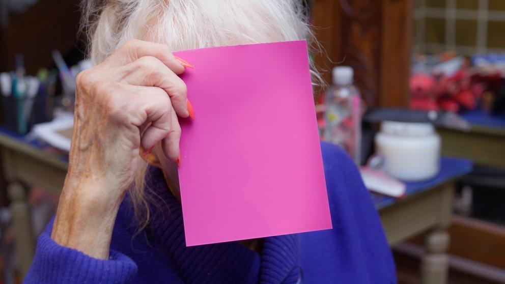 photo of an older woman with platinum blond hair holding up a square of pink in front of her face