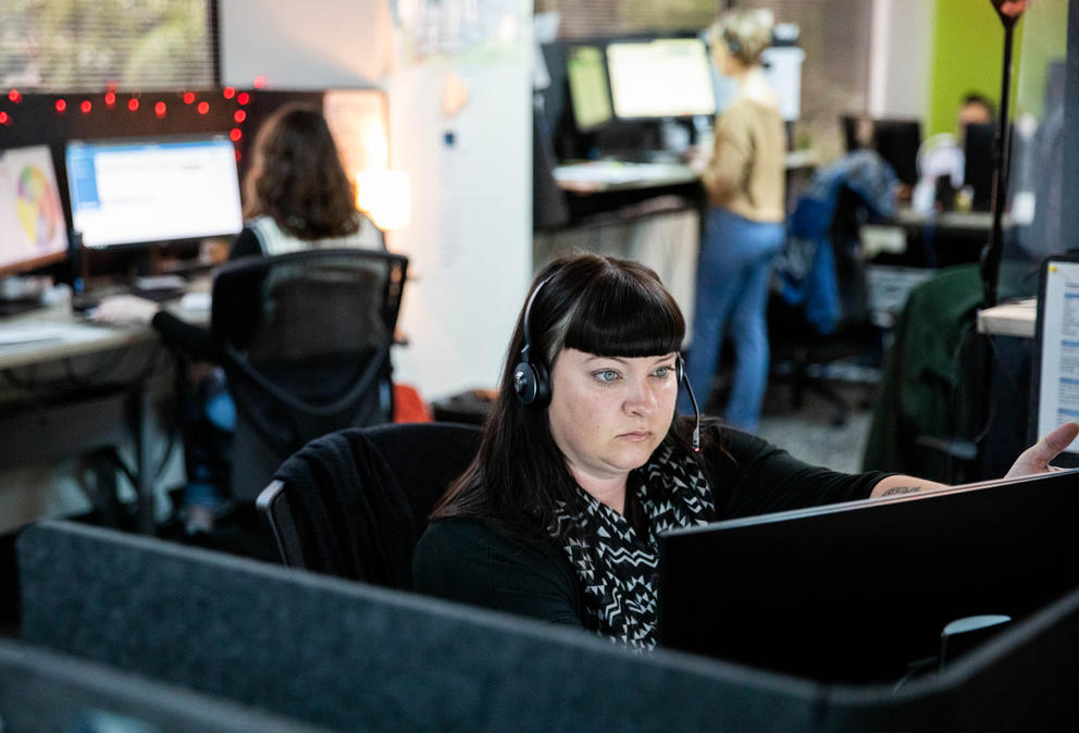 Grove wears a headset and stares intently at a computer screen while other workers are visible in cubicles behind her