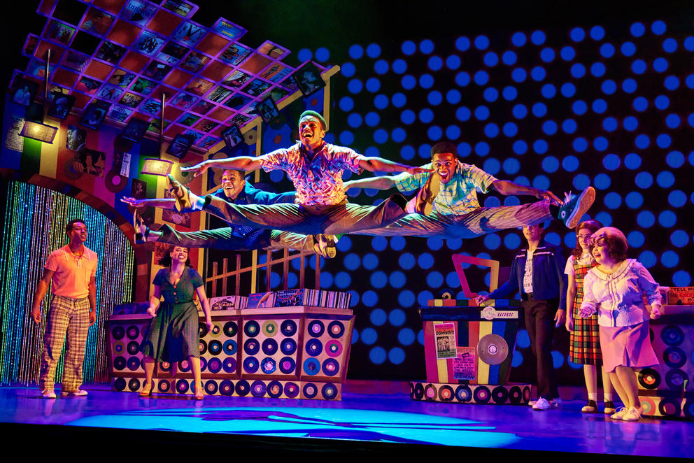 On a colorful stage set, people in brightly patterned outfits do splits in the air as onlookers watch; the lighting is bright hues of purple, blue and pink