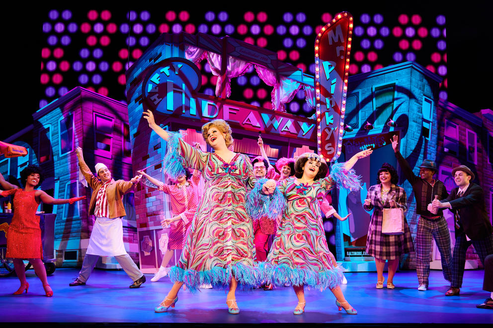 Two women in matching patterned dresses hold hands up and raise the opposite hands upwards on a wildly colored stage