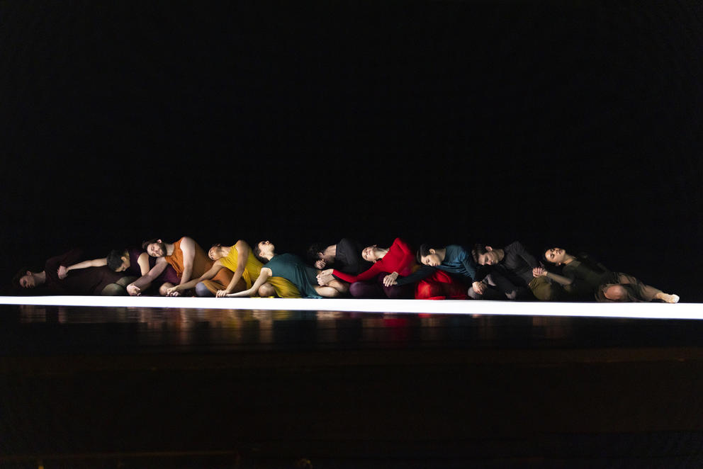 a dark stage in which a row of dancers in rainbow colored costumes sit at the edge of a ray of light