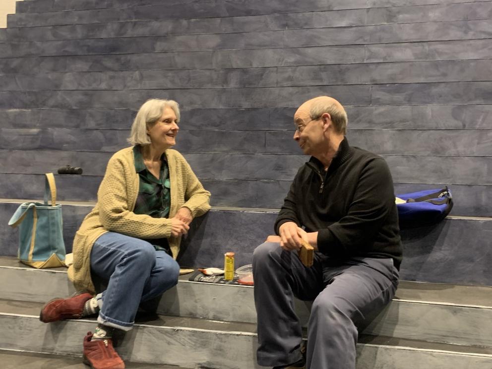 a woman and man in street clothes sit and look at each other in a conversational pose on a gray stage set