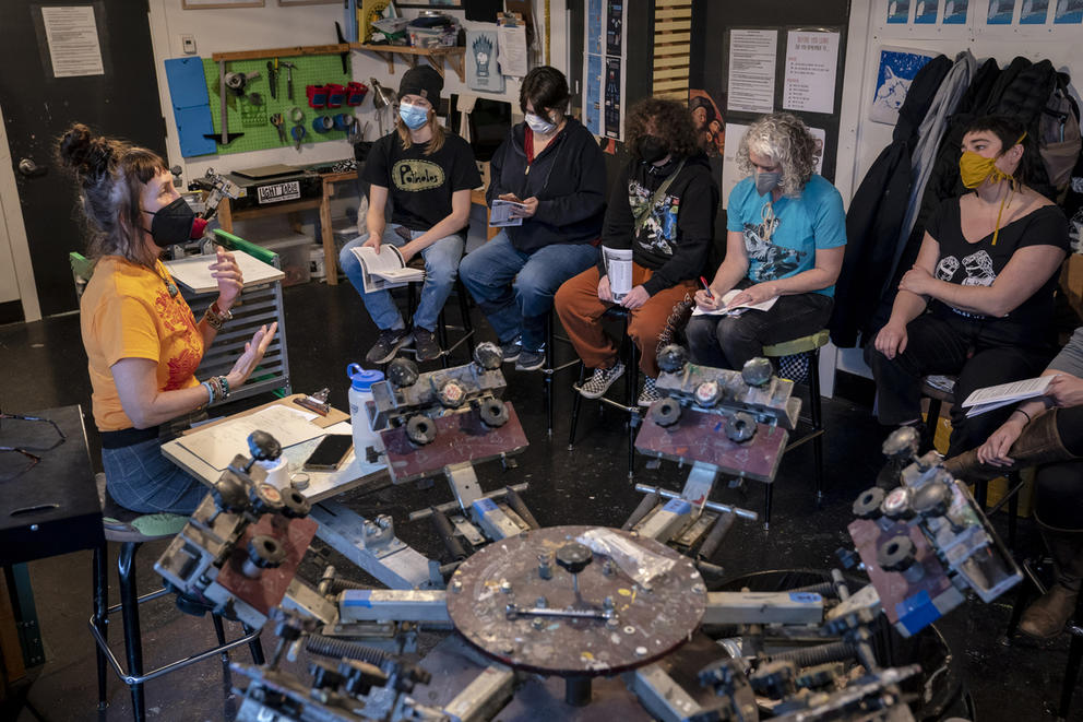 Several people sit in chairs on the right to face a teacher on the left. All are wearing masks and they're in a studio full of different tools and items