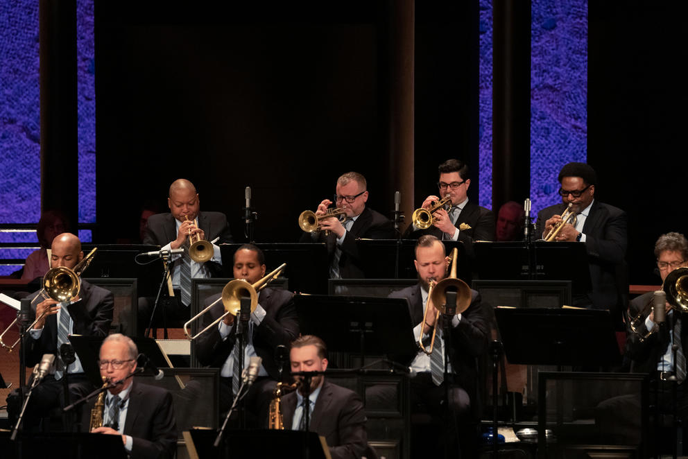 Men sit and play trumpets and trombones on a stage with a purple-lit background