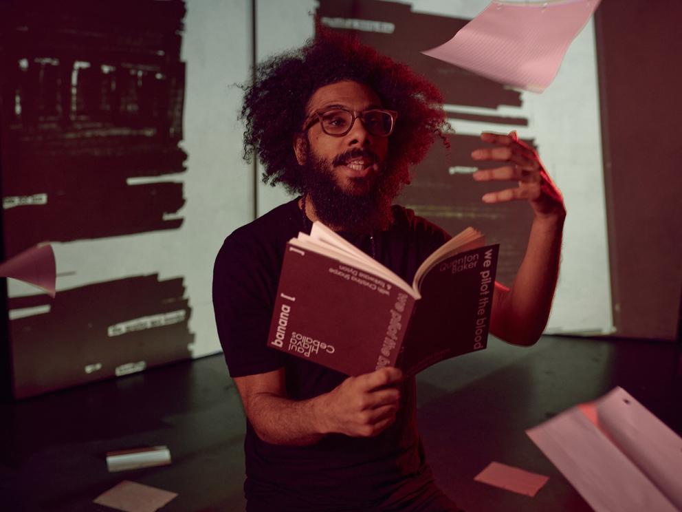 photo of a man in glasses reading aloud from a book while paper cascades around him