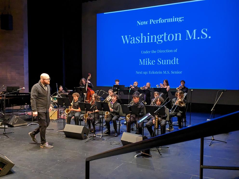 Middle schoolers on stage playing instruments in front of a large blue screen