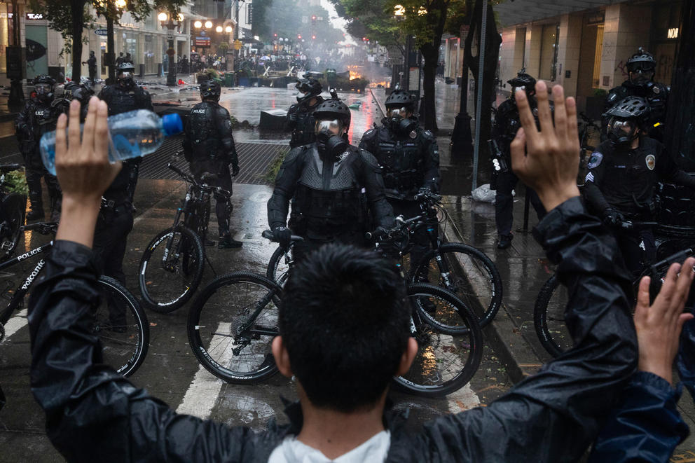 An individual holds his hands over his head while facing a line of bicycle police