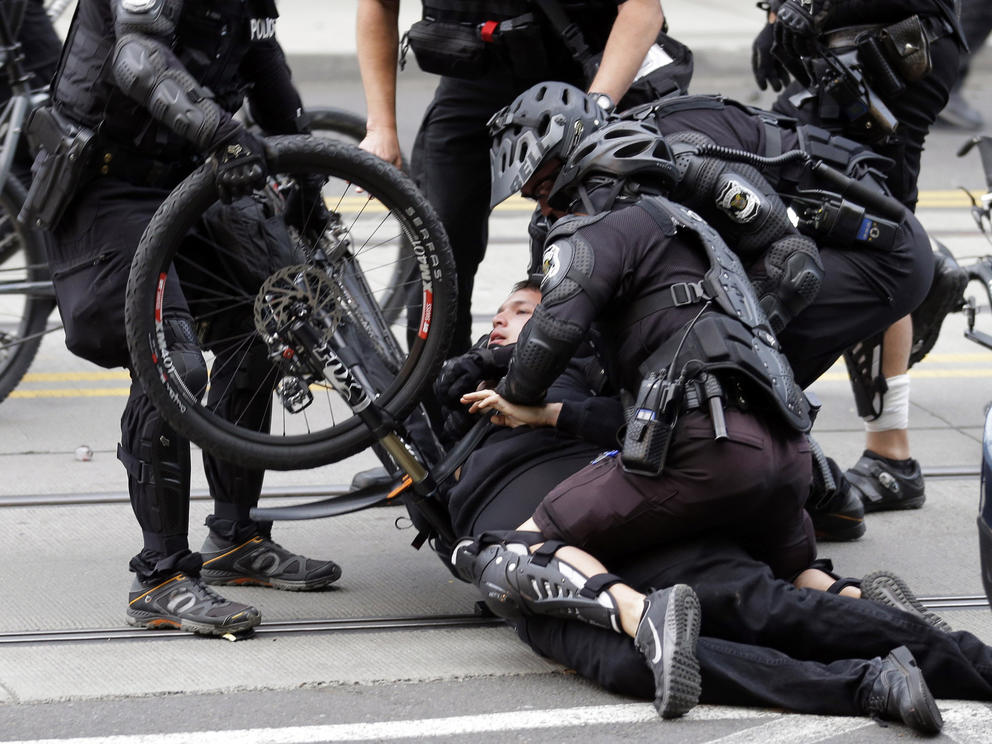 Police in body armor subdue a person using a bike