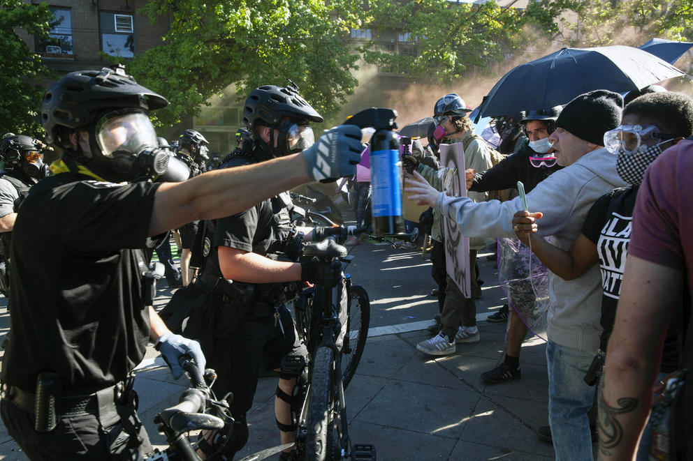 A helmeted officer sprays pepper spray at demonstrators