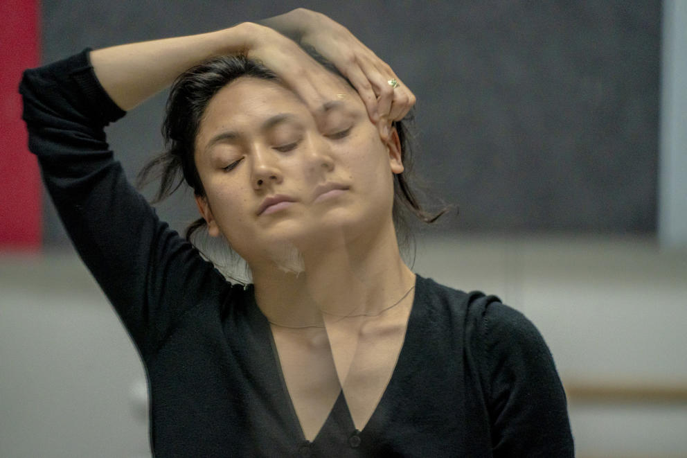 A close up of a dancer in black rehearsing, she has one arm over her head and is seen in double, reflected in a mirror