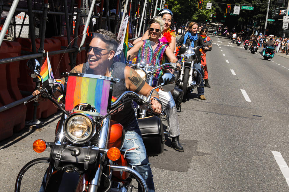 Riders on rainbow covered motorcycles smile as they ride down the street