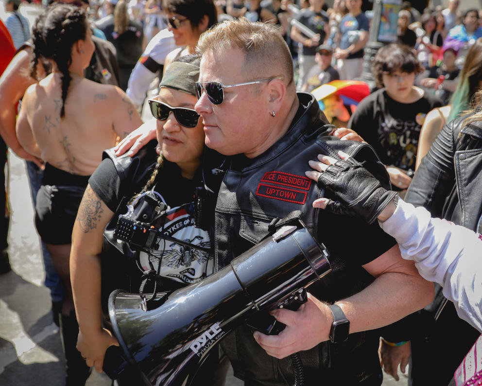Two people wearing leather vests and sunglasses hug each other amid a crowd of people. One holds a megaphone.