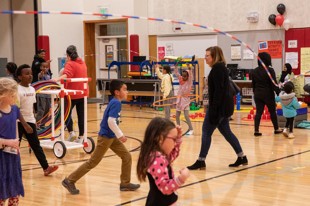 Groups of adults and kids play with jump ropes and hula hoops in a school gym.