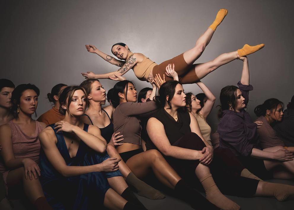 photo of a large group of dancers dressed in brown lifting one dancer over their heads