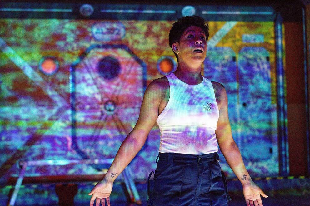 photo of a stage production with an actress in a white tank top with colorful projections across her body