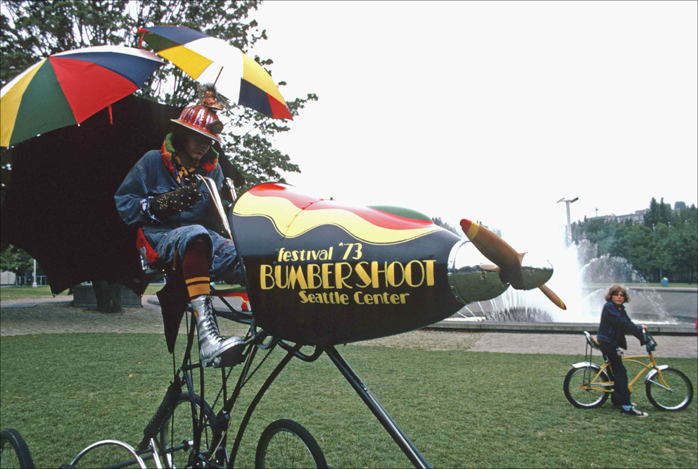 a vintage photo of a clownlike person riding a bike-plane combo with umbrellas attached