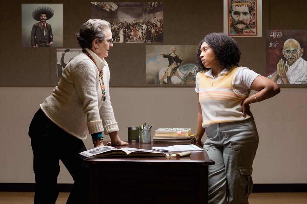 photo of a staged play with two women, one white, one black, facing off over a classroom desk