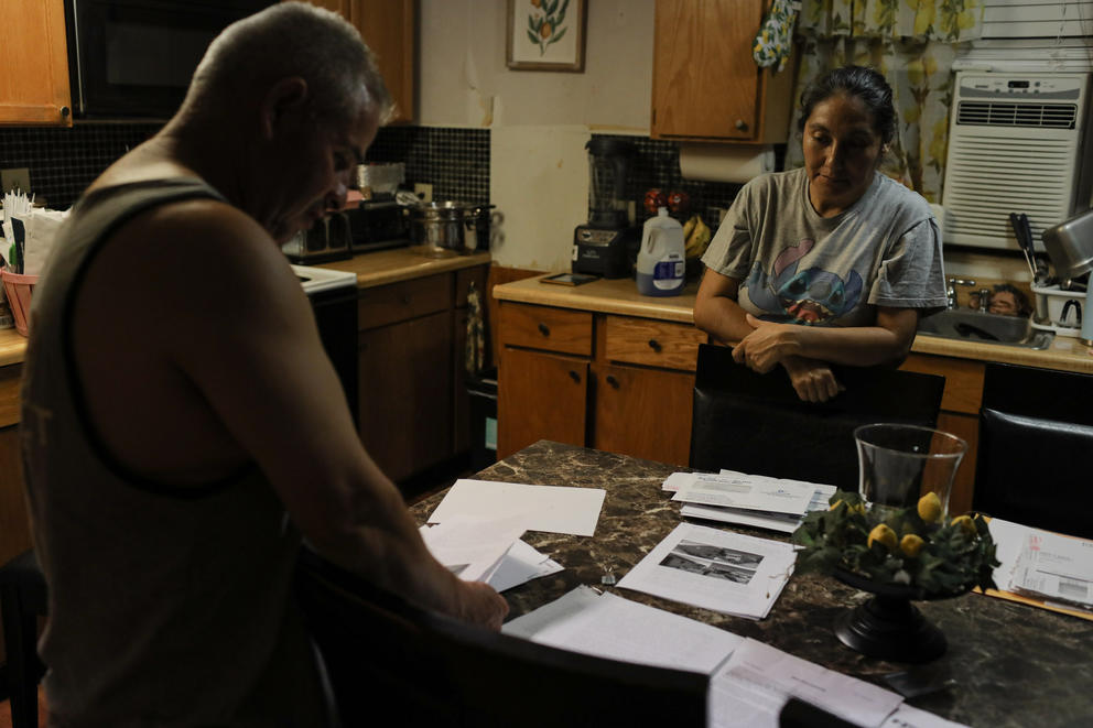 A couple look down at a table with bills and notices on it in their kitchen