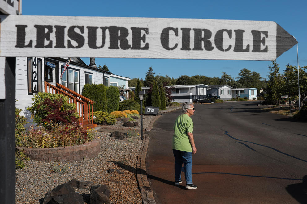 A woman in a bright green shirt walks down a street in a mobile home park, above her there is a street sign that reads Leisure Circle
