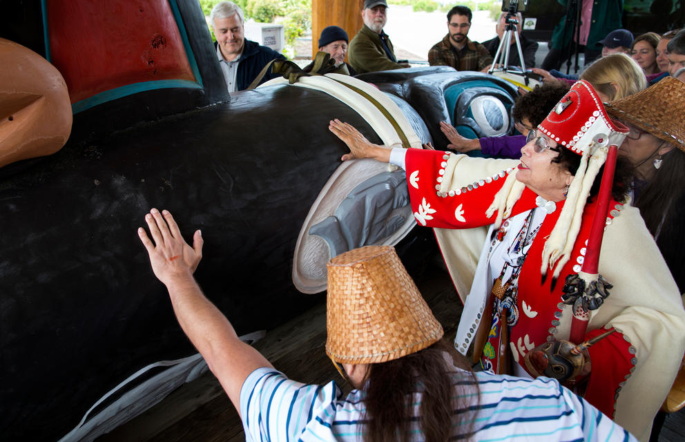 Two people place their hands on a 16-foot totem pole in the shape of an orca.