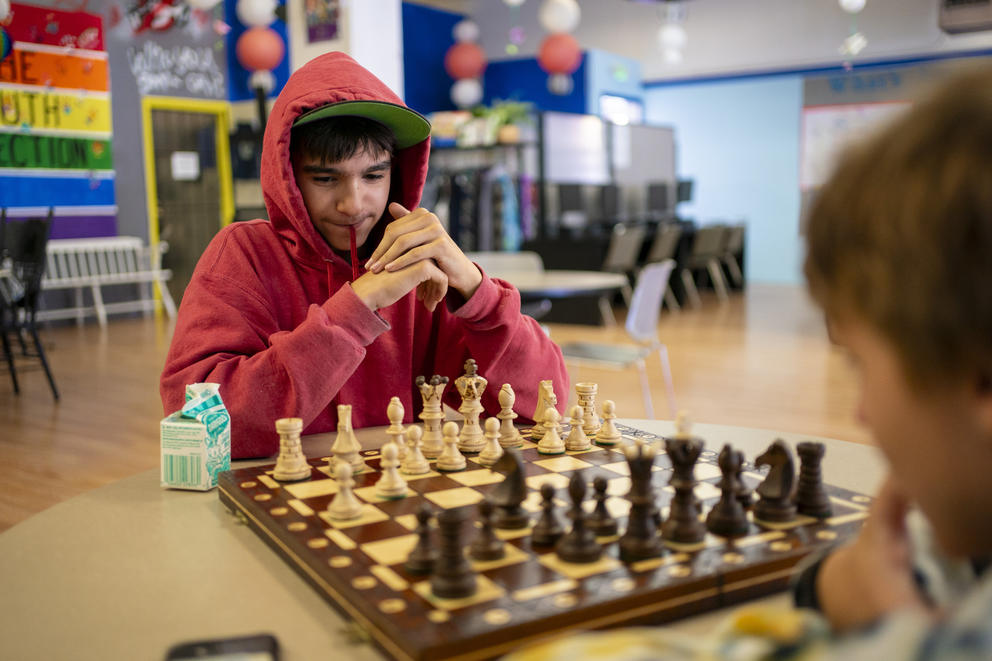 Riley, in a red hoodie sweatshirt, sits looking at a chess board across from another player.