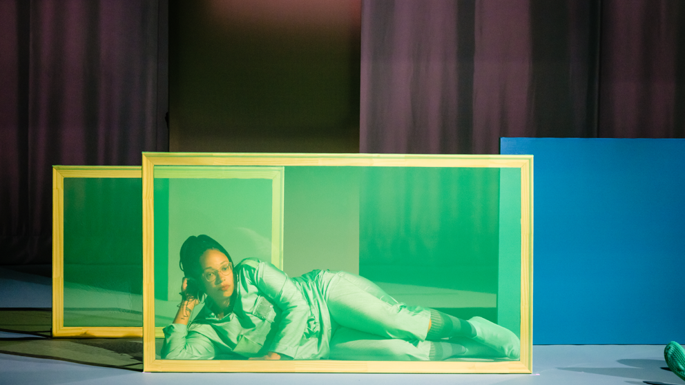 a Black woman lying down on a stage, partially obscured by a green tinted rectangular screen