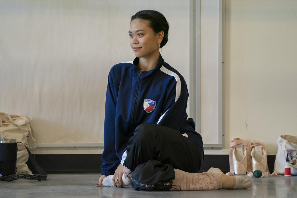 A ballerina half-smiles and stretches her legs on the floor, one over the other