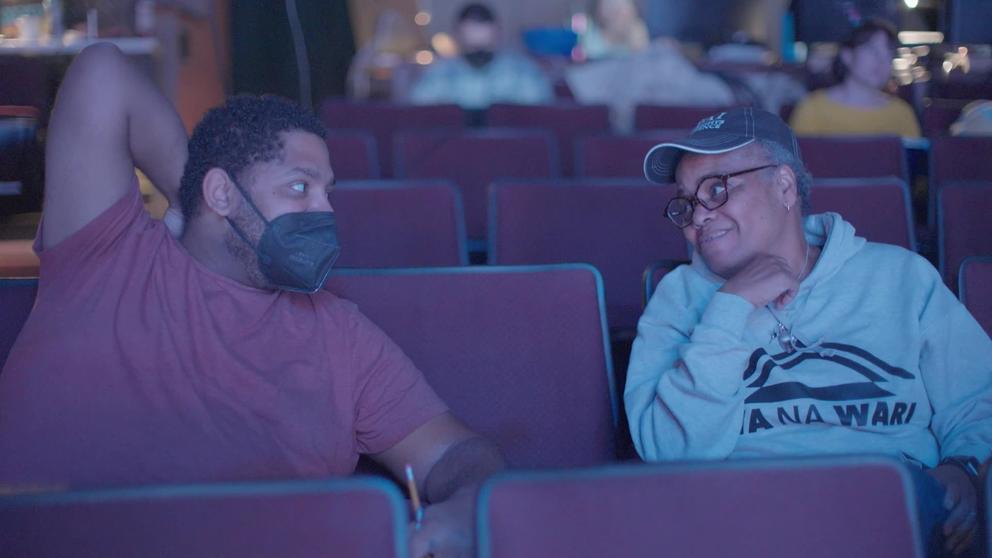 video still of a man and a woman talking in theater seats in a darkened theater