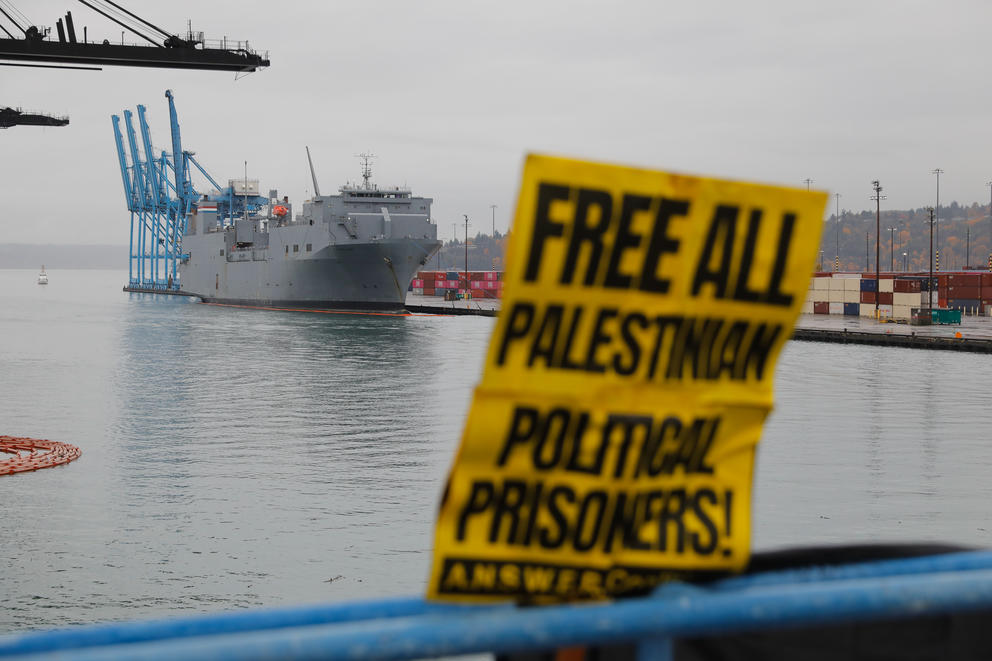 A gray ship sits on a pier, a sign in the foreground reads Free All Palestinian Political Prisoners