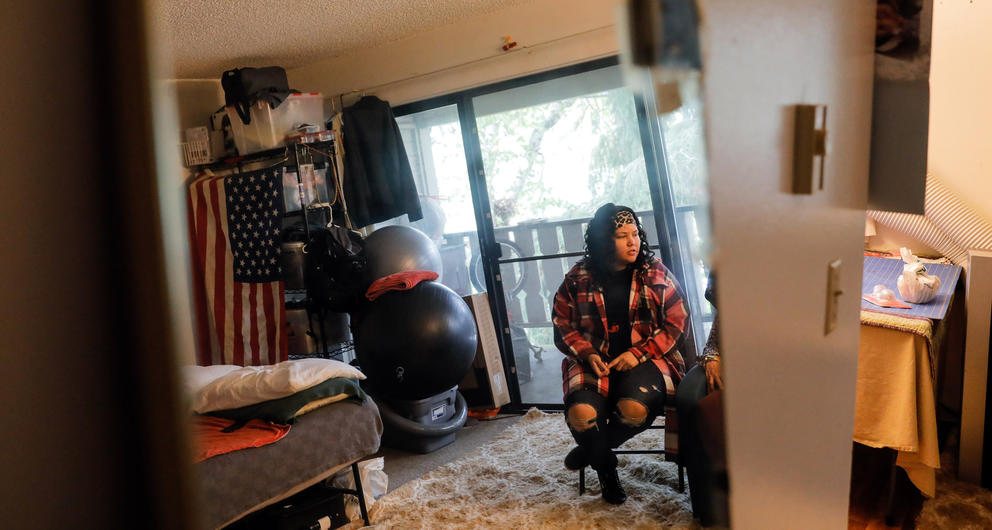 Janell Braxton sits in a folding chair wearing a flannel shirt while reflected in a mirror at her mother's apartment.