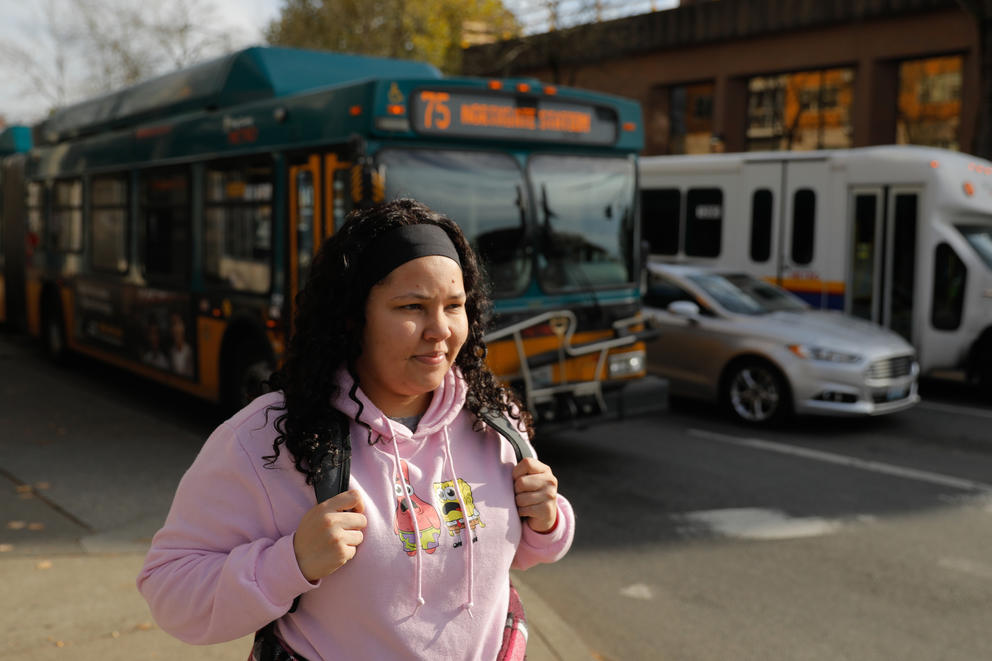 Janell walks along a street with a backpack over her shoulders and a transit bus behind her.