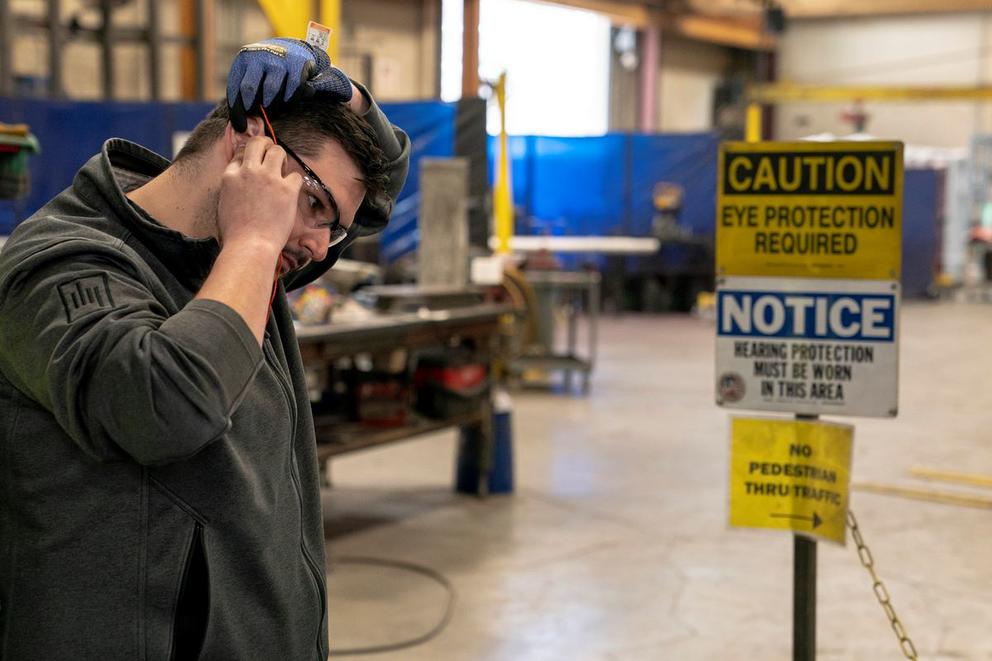 A worker wearing glasses and ear protection with safety signage in the background