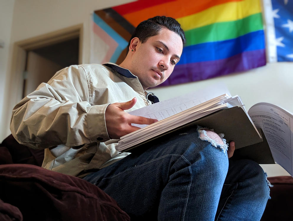 A person sits while reading documents, an LGBTQ+ pride flag is on the wall in the background.