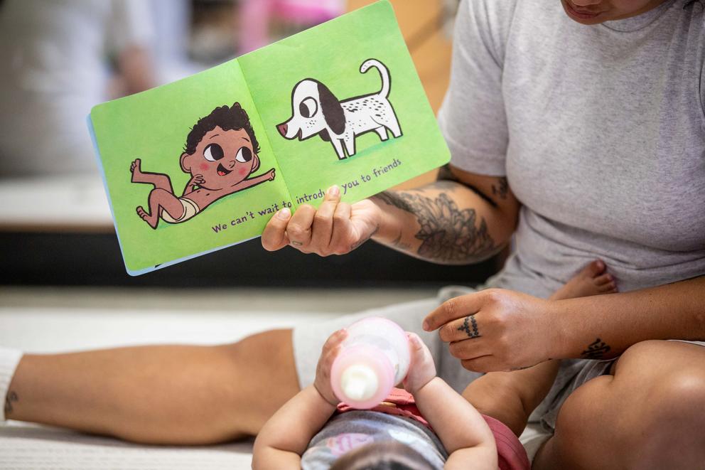 A hand holds up a book on a page that reads "We can't wait to introduce you to new friends" over a baby