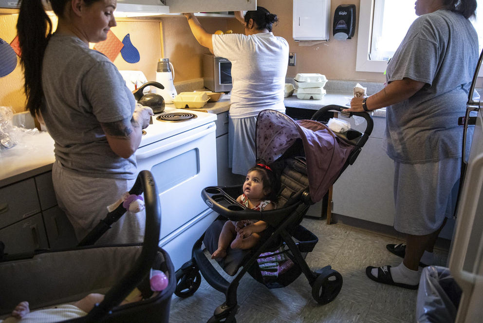 A baby looks up from a stroller in the middle of a kitchen where three women are preparing dinner