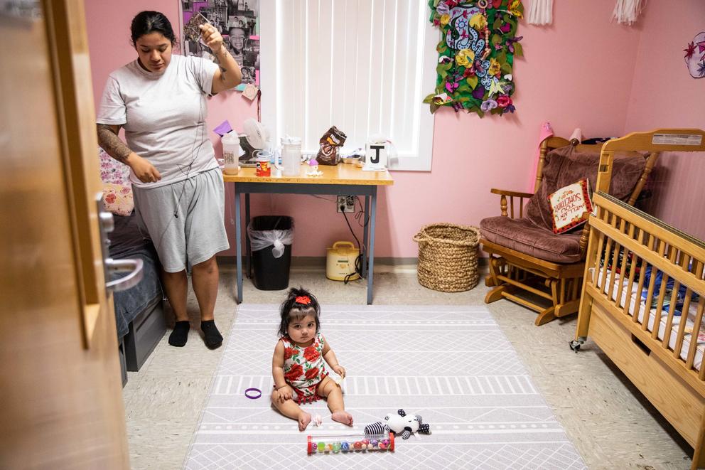 A baby looks up from the floor of a pink bedroom with a bed and a crib. Christina Torres stands to the left. 