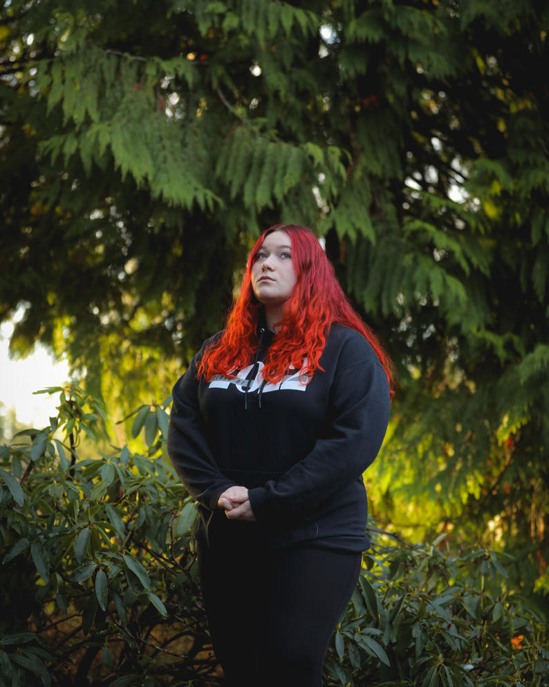 A woman with red hair and a black sweatshirt stands in front of tree