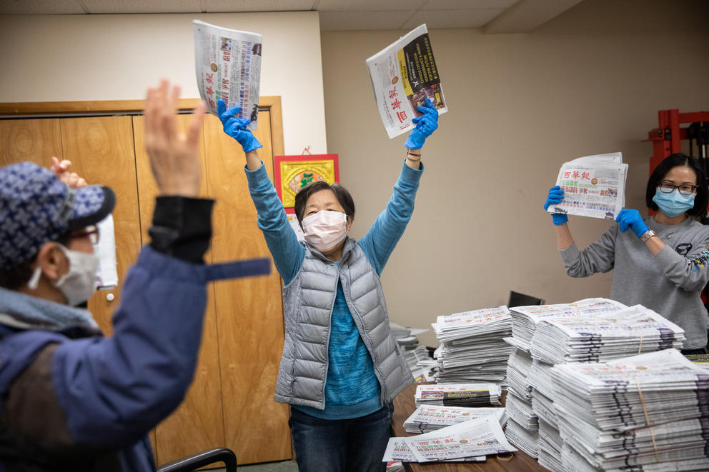 People hold up newspapers from a stack of them on a table