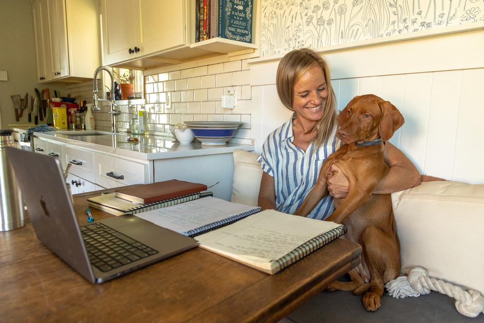 Monica Hines sits at a kitchen table with her dog in front of a laptop and notebook