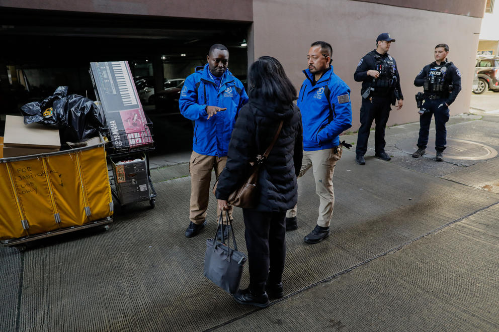 Two police officers stand in the background as two men in blue CARE Team jackets talk to a woman with her back turned in an alleyway