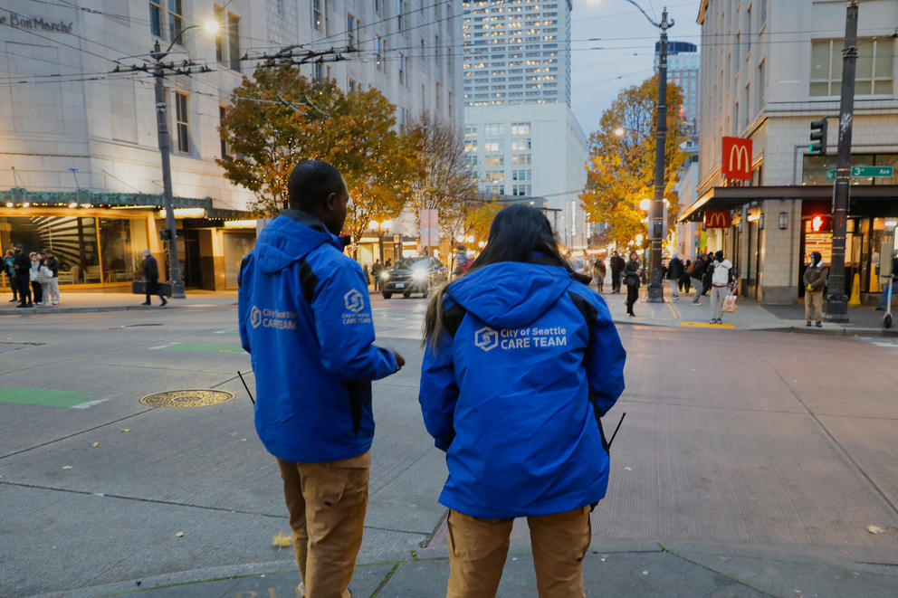 A man and a woman in blue CARE team jackets are seen from behind as they look out across a downtown intersection