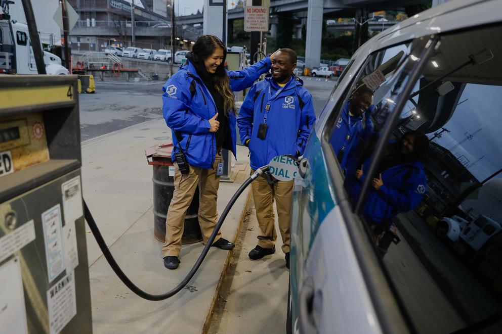 A woman and a man in blue jackets laugh as they pump gas into their car at a gas station